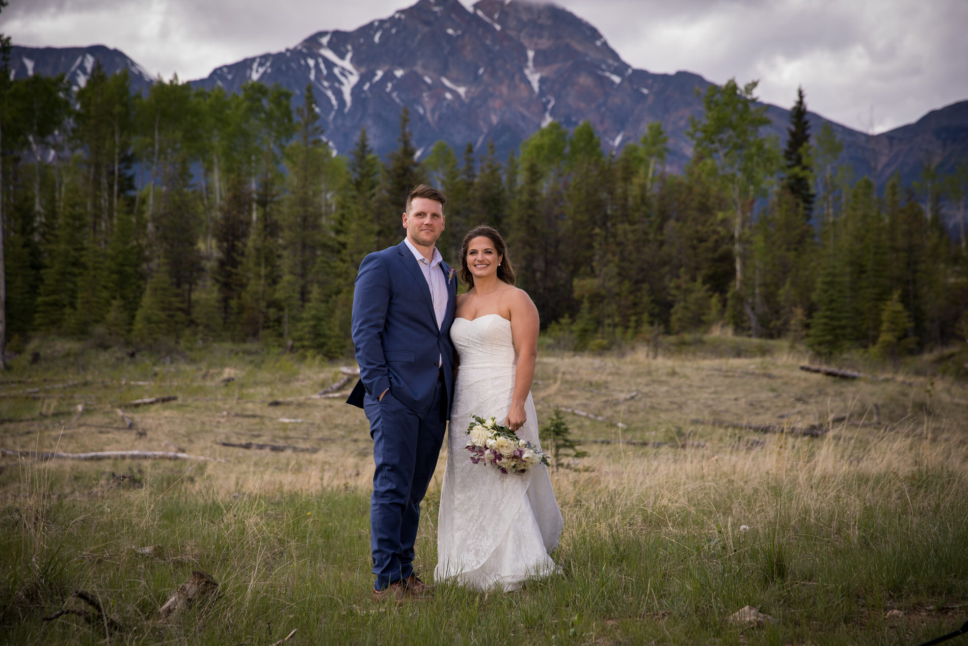 Bride and groom take wedding photos in a field with Pyramid Mountain behind them in Jasper, AB